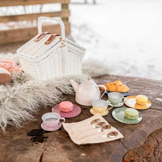 A styled picnic scene outdoors on a rustic wooden table with a white wicker picnic basket, a pink teapot, pastel cups and saucers, macarons, a croissant, and a fur throw blanket, with a snowy background.