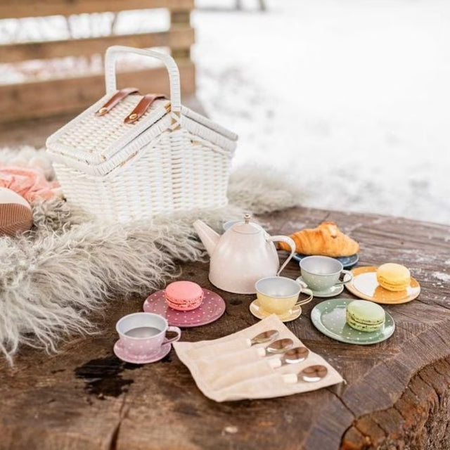A styled picnic scene outdoors on a rustic wooden table with a white wicker picnic basket, a pink teapot, pastel cups and saucers, macarons, a croissant, and a fur throw blanket, with a snowy background.