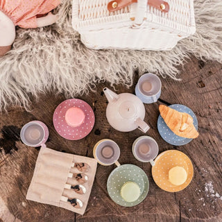 A pastel kids tea party set arranged on a rustic wooden surface with a white wicker basket and faux fur. The set includes a teapot, cups, saucers, a roll of spoons.