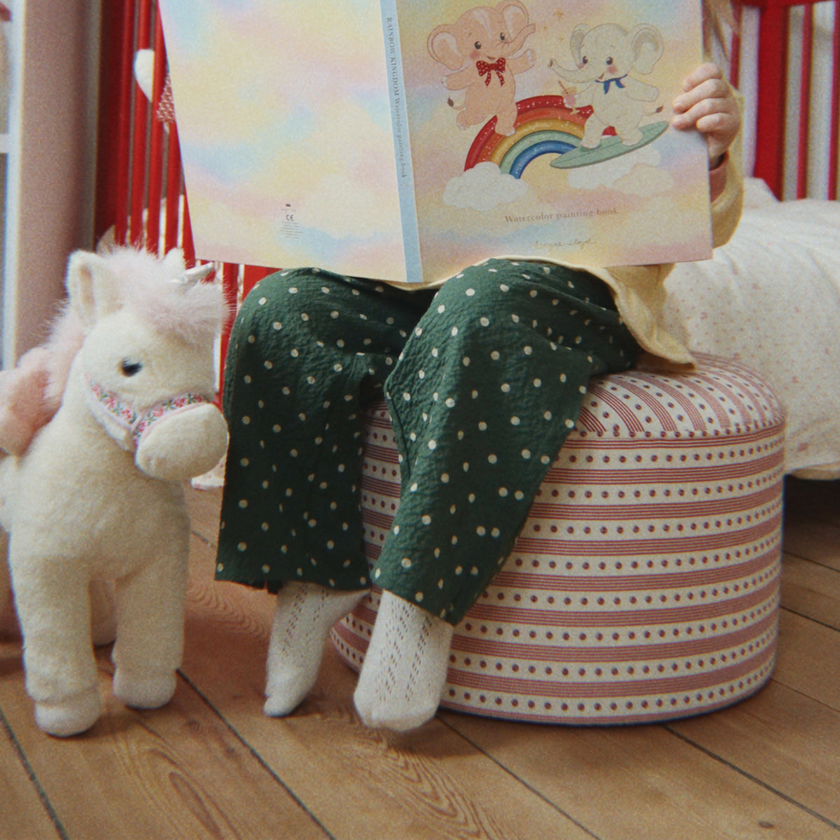 A child sits on a pink and white striped pouf in a nursery, holding an open illustrated book. The child wears dark green polka-dot pajama pants and white socks, with a plush unicorn toy beside them on a wooden floor.