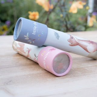 Two children's toy kaleidoscopes labeled 'little dutch' resting on a light wooden surface with a blurred garden background. One is pink with a mesh lens, and the other is white with a blue rim.
