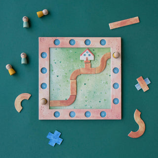 A children's wooden board game set arranged on a dark teal background. The square game board has a pink frame, a light green grassy center, a winding tan path, numbered blue circles around the border, and small house and geometric pieces.