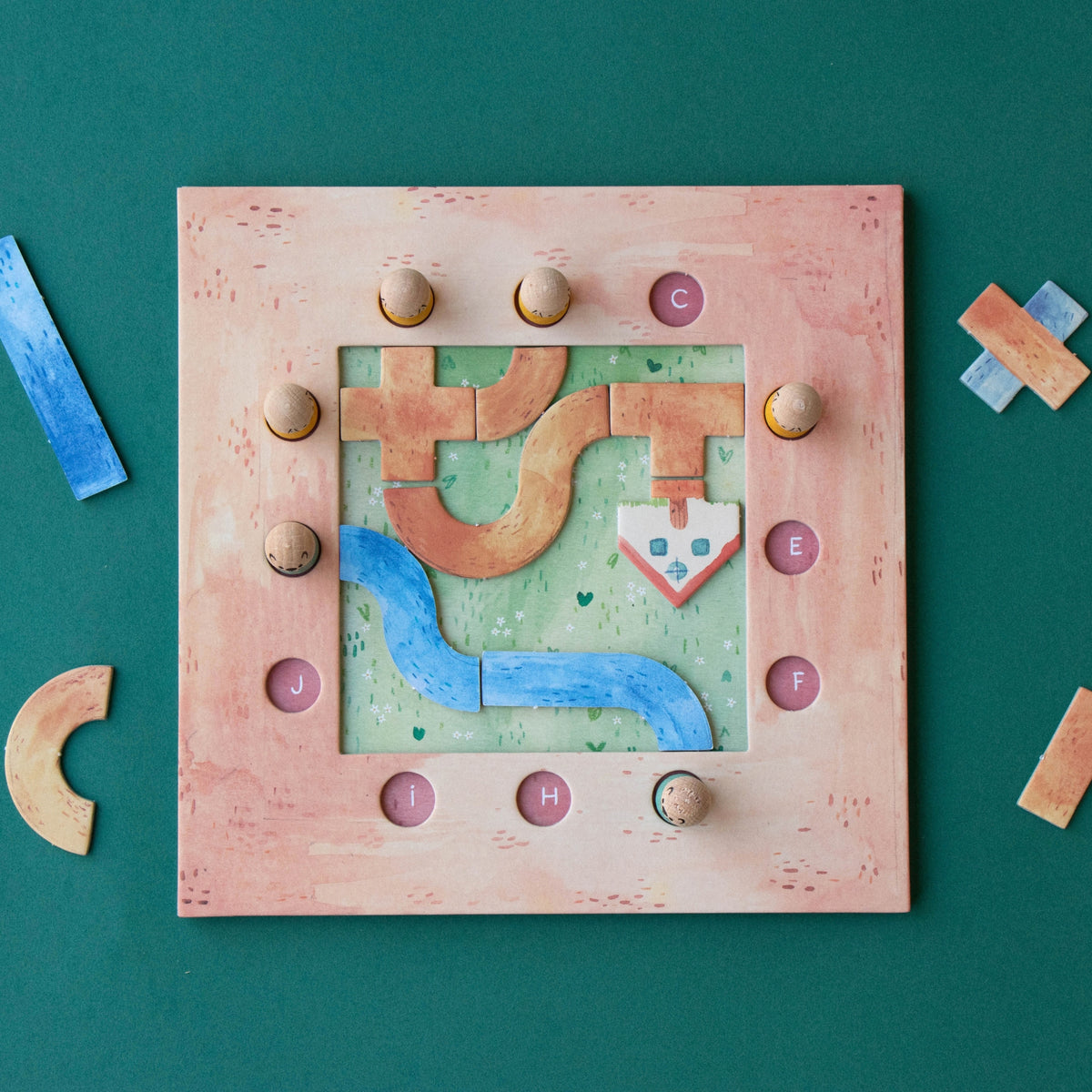 A square wooden puzzle board with a pinkish-beige frame and a colorful geometric maze design, photographed from above on a dark green background with a few loose wooden pieces nearby.