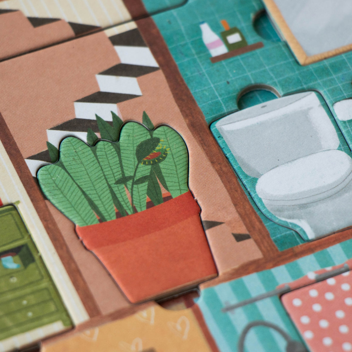 A close-up of a children's cardboard bathroom scene with a green potted plant in an orange pot, a teal bathroom tile with a toilet cutout, and other kitchen props.