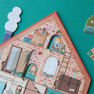 A close-up of a house-shaped puzzle with illustrated interior scenes, including windows, a ladder, suitcase, and small cutout pieces, arranged on a teal background.