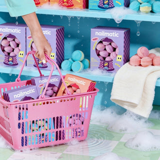 A pink plastic shopping basket filled with Nailmatic mini bath bomb boxes, held by a person's hand in front of shelves of more bath bomb boxes.
