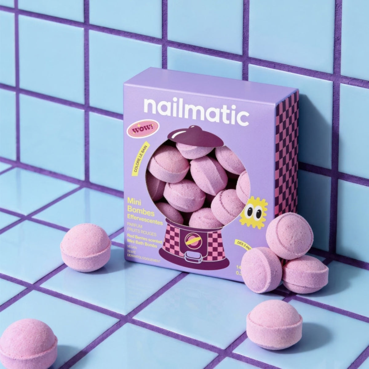 A purple box of nailmatic mini bath bombs sits on a light blue tiled surface with several purple bath bombs scattered around it.
