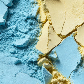Close-up of two textured powder products, one in light blue and one in pale yellow, with cracked, pressed solid pieces layered over loose powders.