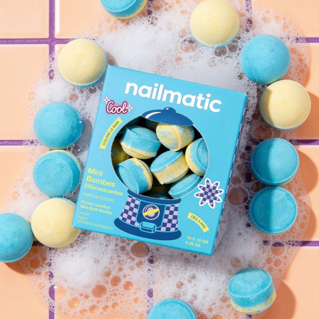 A box of nailmatic mini bath bombs surrounded by blue and pale yellow bath bombs on a peach tiled surface with white bubbly foam.