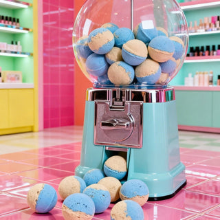 A retro-style turquoise gumball machine filled with blue and beige bath bombs, with several macarons spilled onto the pink tiled floor in a brightly lit candy store.