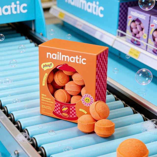 An orange box of naimatic mini bath bombs sits on a blue conveyor belt in a retail setting, with several round orange bath bombs placed in front of the box.