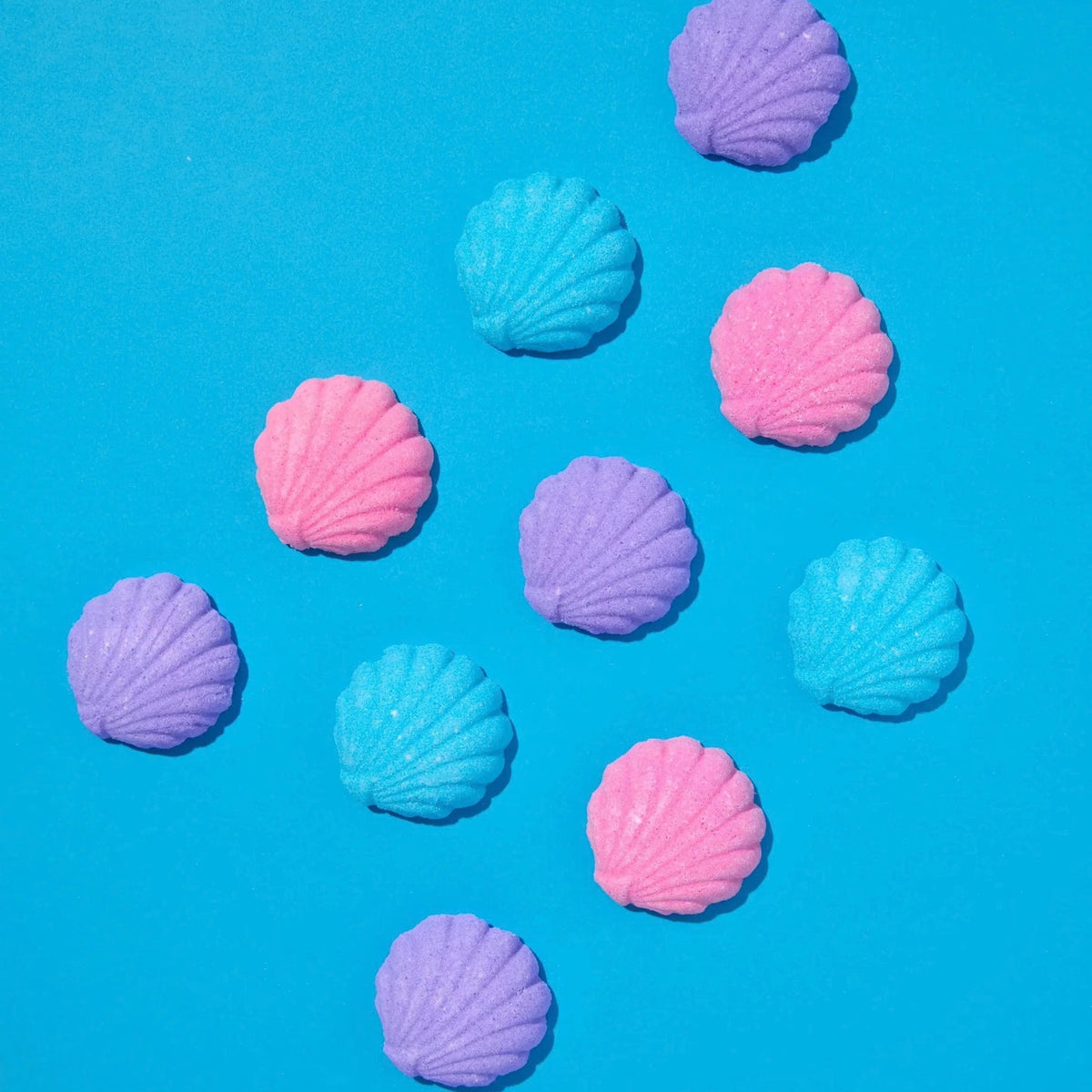 Ten pastel-colored seashell-shaped bath bombs in pink, purple, and light blue, scattered on a bright blue background.