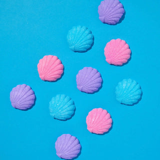 Ten pastel-colored seashell-shaped bath bombs in pink, purple, and light blue, scattered on a bright blue background.