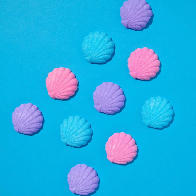 Ten pastel-colored seashell-shaped bath bombs in pink, purple, and light blue, scattered on a bright blue background.