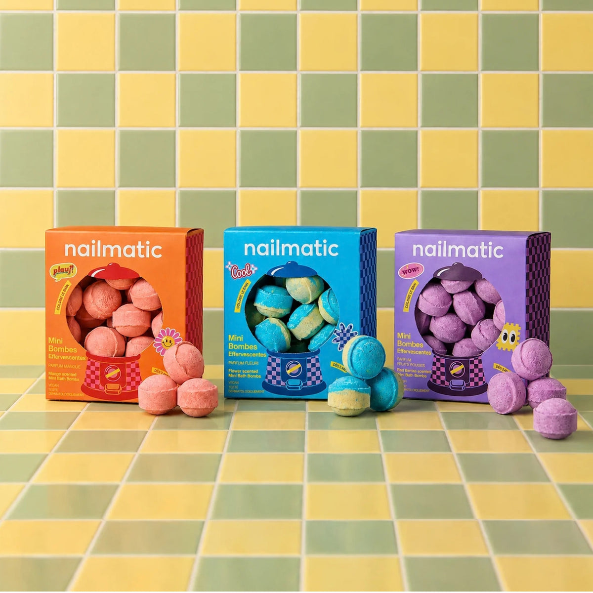 Three colorful nailmatic Mini Bath Bombs boxes lined up on a green and yellow checkered tile surface, with bath bombs spilling out in front of each box.