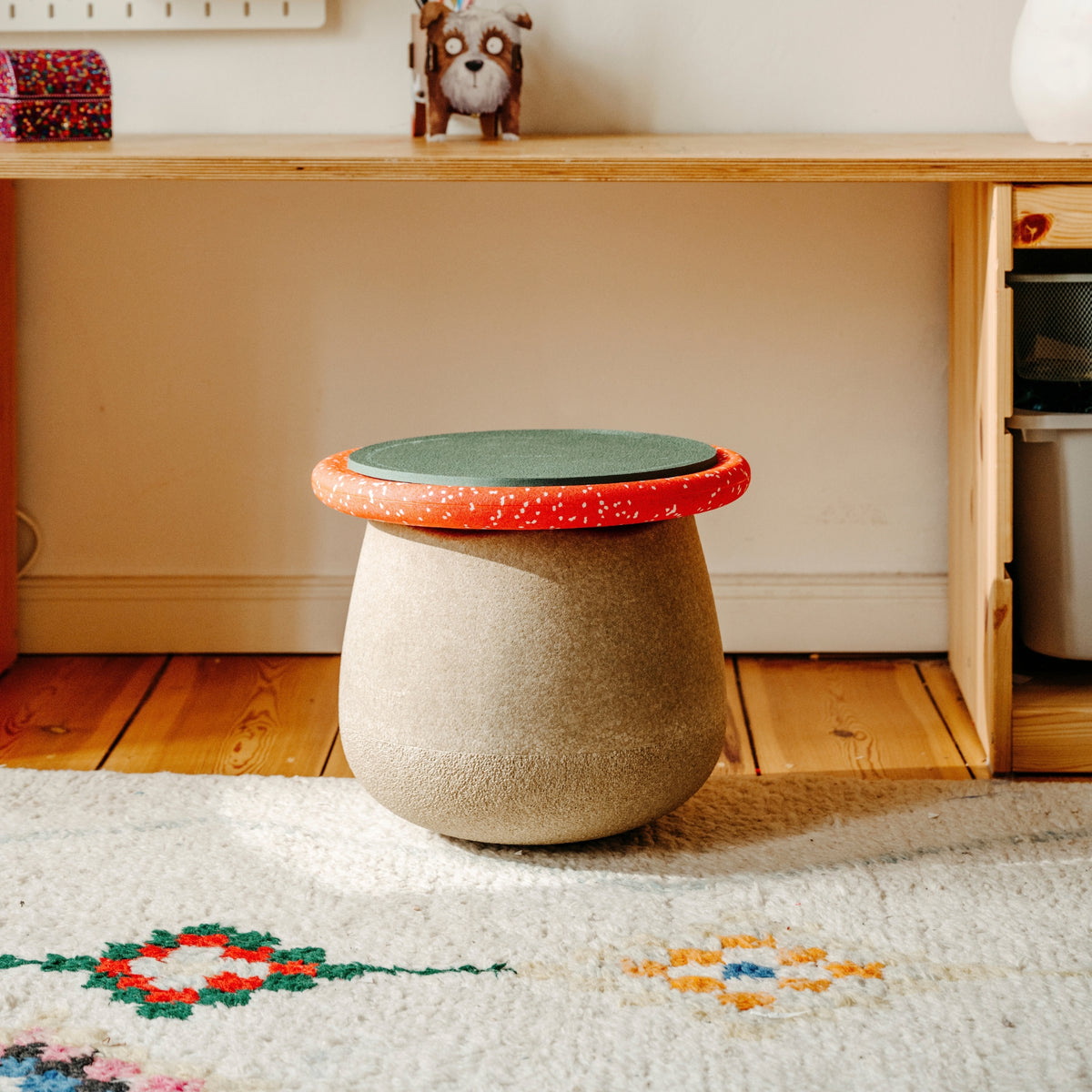 A small round stool with a textured beige body and a lid featuring a green seat pad and red speckled rim, placed on a patterned rug in front of a wooden desk.