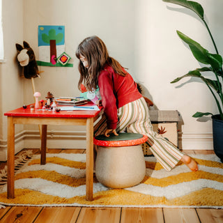 A child in a red top and striped trousers sits on a toadstool-shaped stool next to a small wooden table with a red top in a cosy, sunlit room.