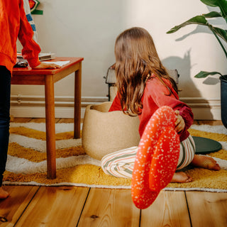 A child sits on a striped rug in a warm indoor room, holding up a bright red speckled round wobble board. Another child in an orange top is partially visible at the left edge, and a wooden table, beanbag, plant, and wood floor are visible in the scene.