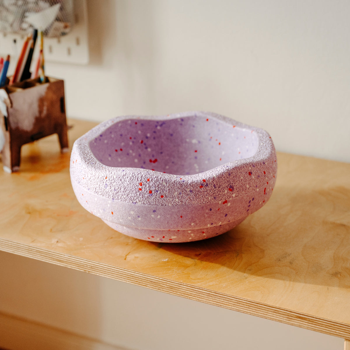 A lavender purple textured Original Stapelstein piece with red and white speckles, sitting on a light wood desk in a home office setting.
