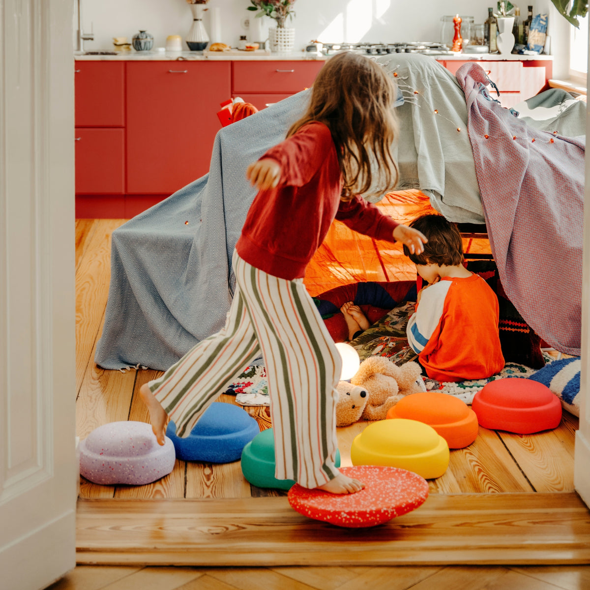 A little girl in a red sweatshirt and striped trousers stands barefoot on a red circular board while another child sits inside a play tent made from blankets, in a bright room with wooden floors and orange cabinets.