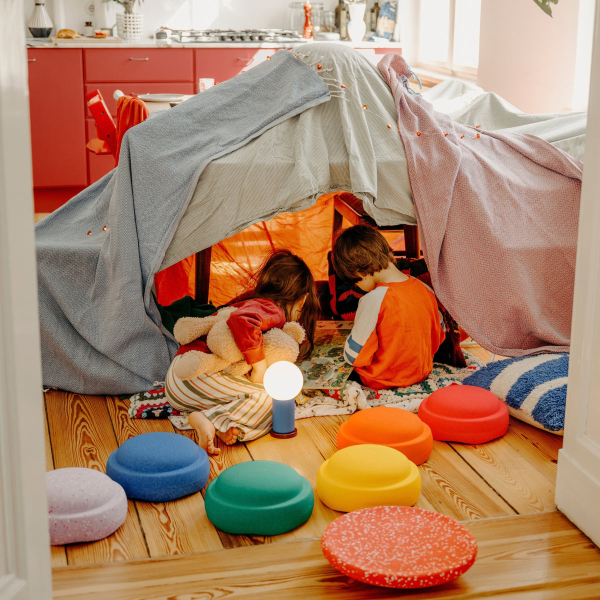 Two children play inside a fabric tent on a wooden floor, with colorful Original Stapelstein pieces arranged around them in a bright room.
