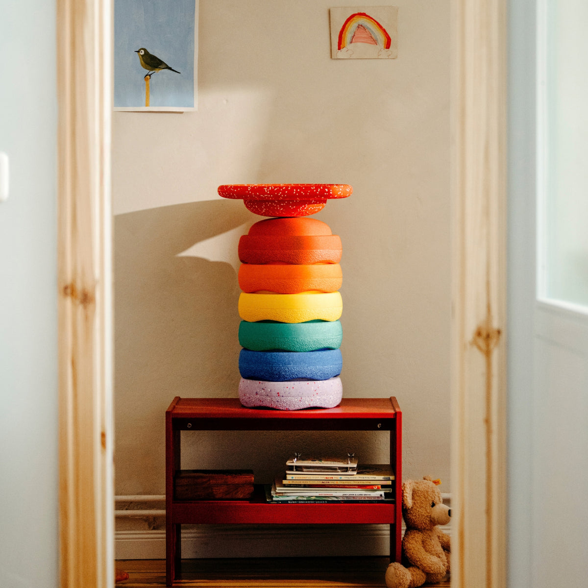A stack of 6 Original Stapelstein pieces in rainbow order with a red speckled board on top, displayed on a small red shelf in a child's room.