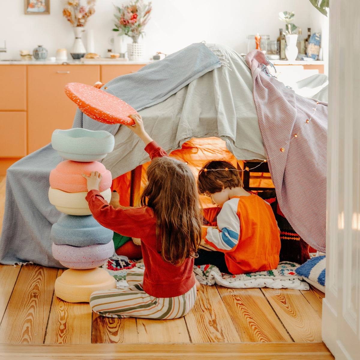 Two children playing inside a cosy fabric play tent on a wooden floor, with one child stacking pastel-colored Original Stapelstein pieces and the other sitting inside the tent. The room has warm orange cabinets and decorative items in the background.