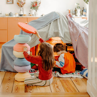 Two children playing inside a cosy fabric play tent on a wooden floor, with one child stacking pastel-colored Original Stapelstein pieces and the other sitting inside the tent. The room has warm orange cabinets and decorative items in the background.