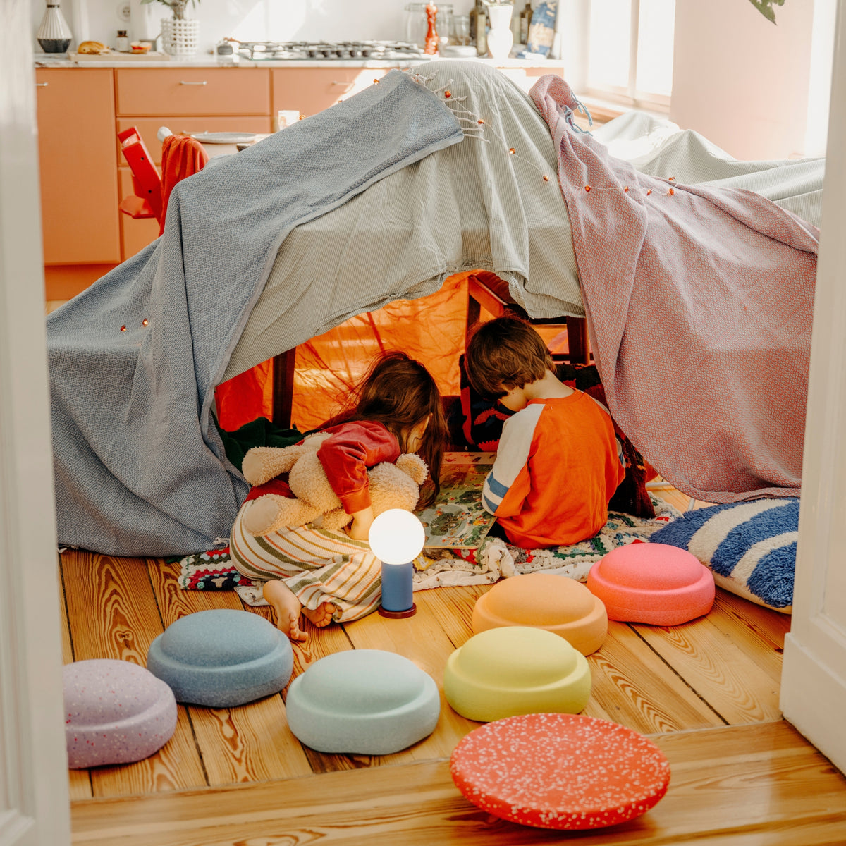 A child's play tent made from draped blankets set up on a wooden floor in a cosy room. Two children are sitting inside the tent whilst their Original Staplestein pieces are placed in path formation outside the tent.