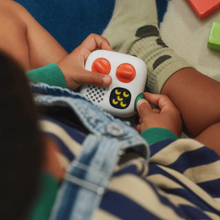 A close-up lifestyle shot of a child's hands holding a Yoto Mini Player 4th Gen with orange dials and a green button while wearing a blue and beige striped shirt and green socks.